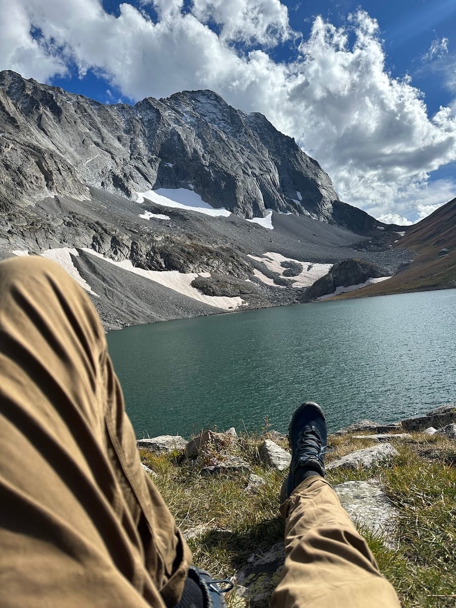 Hiker's legs resting by an alpine lake on the Capitol Peak Trail.