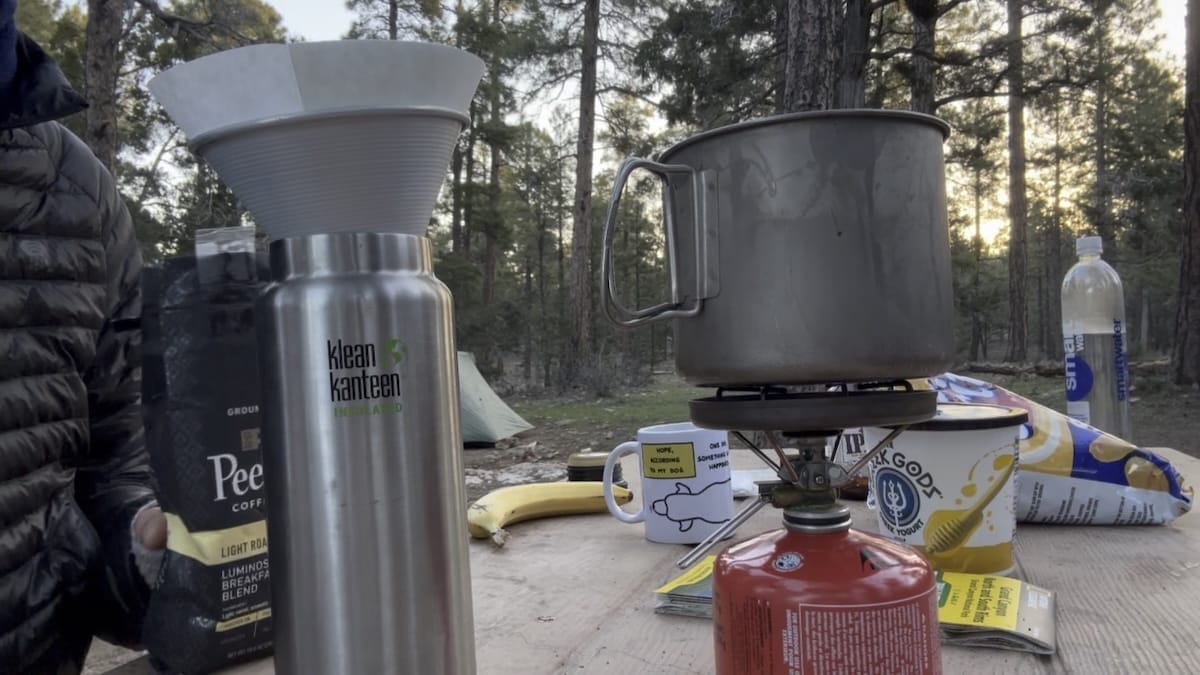 Camping coffee setup at Mather Campground in Grand Canyon with a Klean Kanteen bottle, pour-over filter, titanium kettle on a gas stove, and 'Peet Coffee' bag.