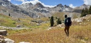 hiker-alpine-meadow-large-snow-capped-peaks Hiker walking into alpine meadow with large snow capped peaks in the distance.