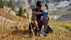 Hiker filtering water with a Sawyer Squeeze sitting on his backpack in a meadow.